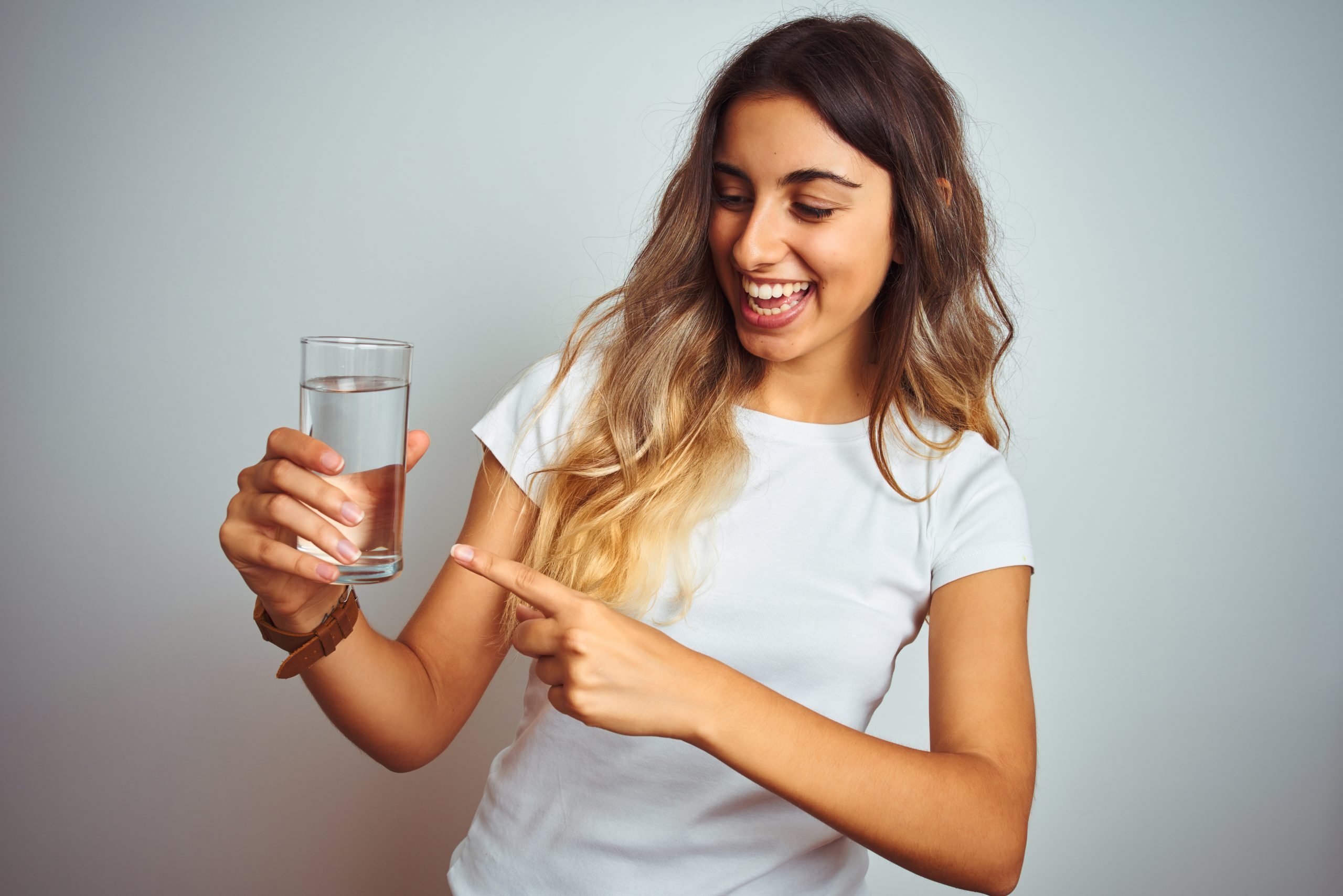 Young beautiful woman drinking a glass of water over white isolated background very happy pointing with hand and finger
